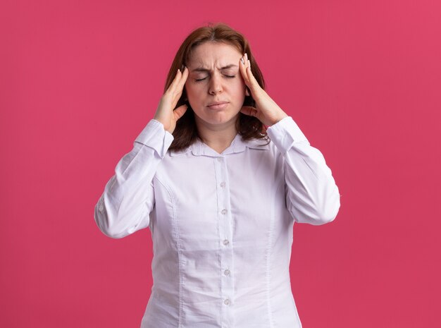 Young woman in white shirt looking unwell touching her temples suffering from headache standing over pink wall