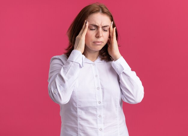 Young woman in white shirt looking unwell touching her temples suffering from headache standing over pink wall