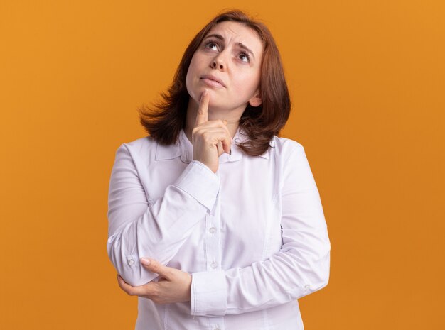 Young woman in white shirt looking up puzzled standing over orange wall