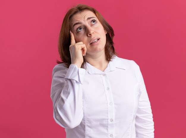 Young woman in white shirt looking up puzzled standing over pink wall