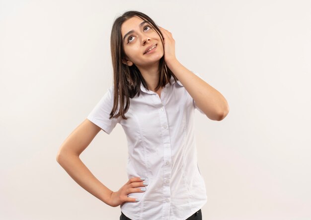 白い壁の上に立っている物思いにふける表情で顔の近くの手で見上げる白いシャツの若い女性