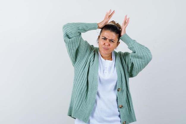 Free photo young woman in white shirt and mint green cardigan raising hands above head, grimacing and looking harried