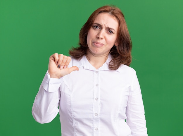 Foto gratuita giovane donna in camicia bianca che punta a se stessa guardando fiducioso in piedi oltre il muro verde