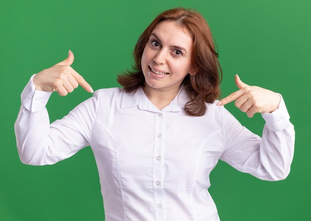 Young woman in white shirt pointing at herself smiling confident standing over green wall