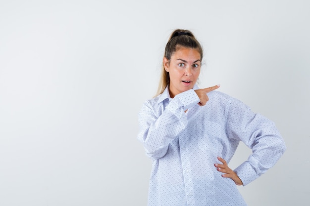 Young woman in white shirt pointing right with index finger, holding hand on hip and looking surprised