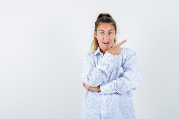 Young woman in white shirt pointing right with index finger and looking surprised