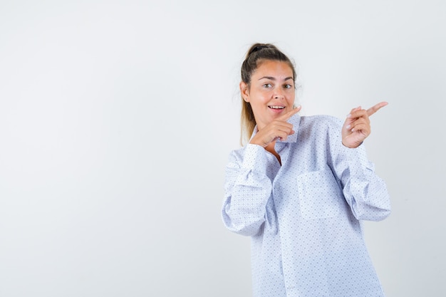 Молодая женщина в белой рубашке указывает вправо указательными пальцами и выглядит счастливой