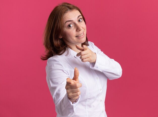 Young woman in white shirt pointing with index fingers at front smiling confident standing over pink wall