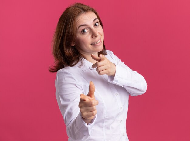 Молодая женщина в белой рубашке, указывая указательными пальцами вперед, уверенно улыбаясь, стоит над розовой стеной