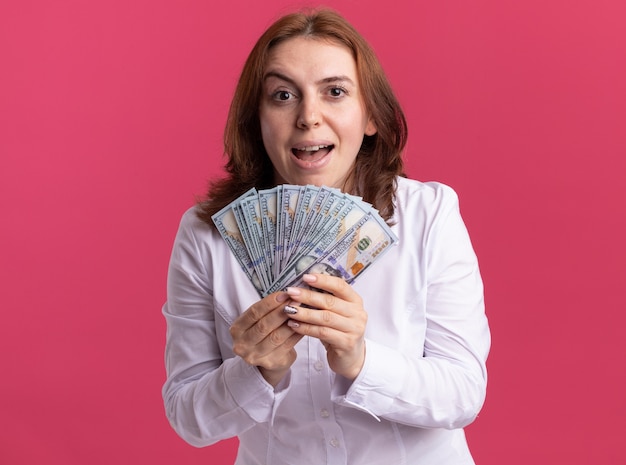 분홍색 벽 위에 서 행복하고 흥분된 전면을보고 현금을 보여주는 흰 셔츠에 젊은 여자