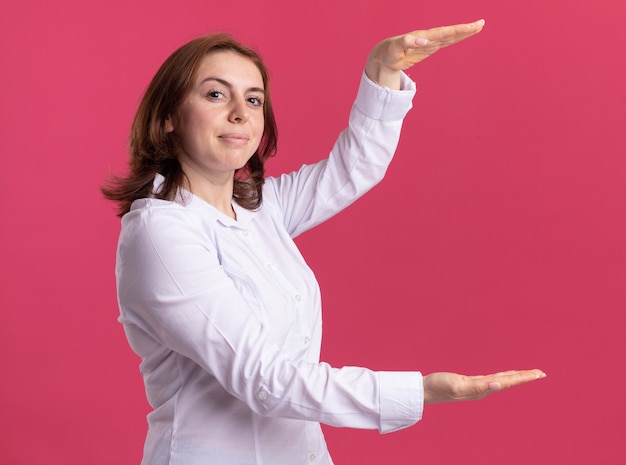 Молодая женщина в белой рубашке, показывающая размерный жест руками, уверенно улыбаясь, измеряет символ, стоя над розовой стеной