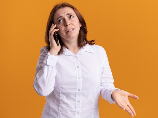 Young woman in white shirt talking on mobile phone being confused standing over orange wall