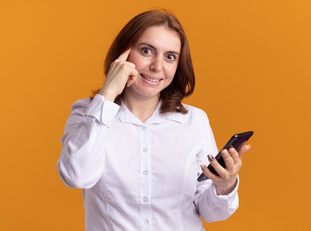 Young woman in white shirt with smartphone looking at front smiling pointing with index finger at her temple standing over orange wall