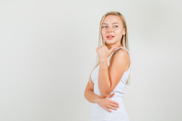 Young woman in white singlet pointing to side with thumb and looking confident