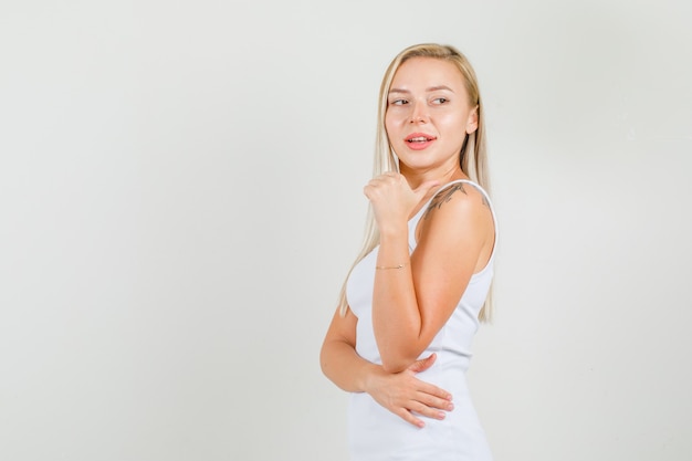 Young woman in white singlet pointing to side with thumb and looking confident