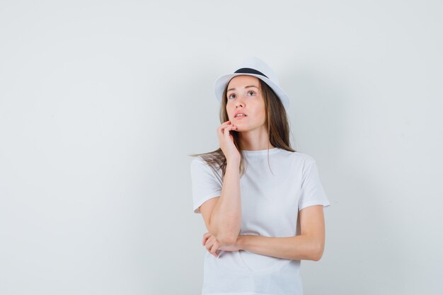 Young woman in white t-shirt, hat looking up and looking pensive.