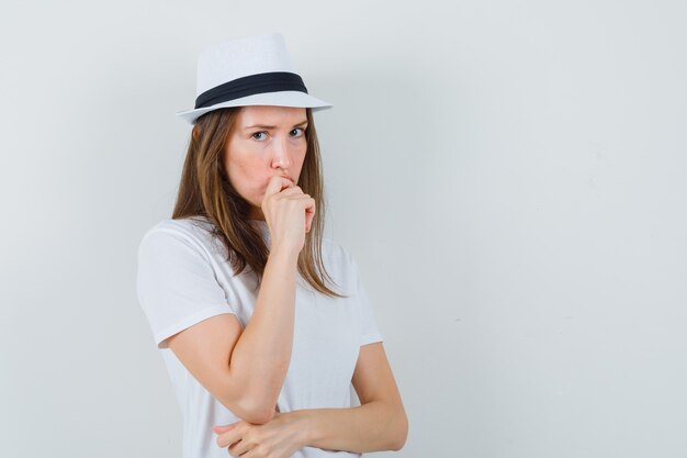 Молодая женщина в белой футболке, шляпе, стоящей в позе мышления и выглядящей обеспокоенной.