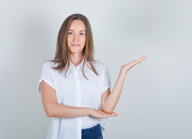 Young woman in white t-shirt, jeans welcoming or showing something and looking glad