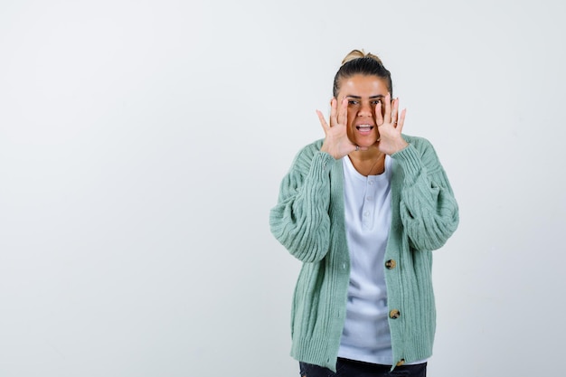 Foto gratuita giovane donna in maglietta bianca e cardigan verde menta che si tiene per mano vicino alla bocca mentre chiama qualcuno e sembra felice