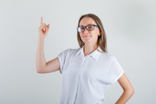 Молодая женщина в белой футболке показывает пальцем вверх и выглядит веселой