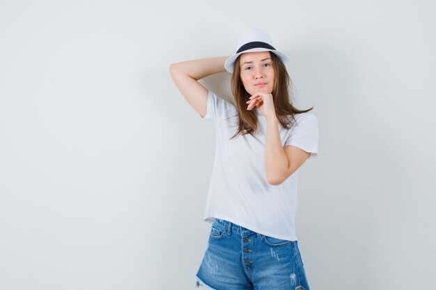 Young woman in white t-shirt, shorts, hat posing while standing and looking delightful.