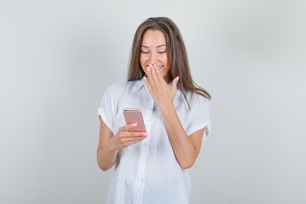 Young woman in white t-shirt using smartphone with hand on mouth and looking happy