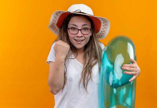 Young woman in white t-shirt wearing summer hat  holding inflatable ring clenching fist looking excited and happy standing over orange wall