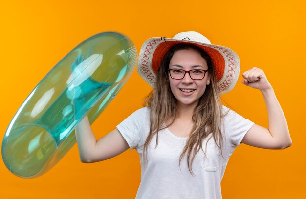 Young woman in white t-shirt wearing summer hat  holding inflatable ring clenching fist looking excited and happy standing over orange wall