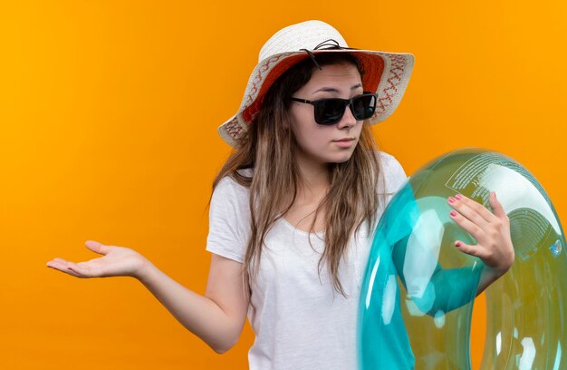 Young woman in white t-shirt wearing summer hat  holding inflatable ring looking confused and uncertain spreading palms to the side standing over orange wall