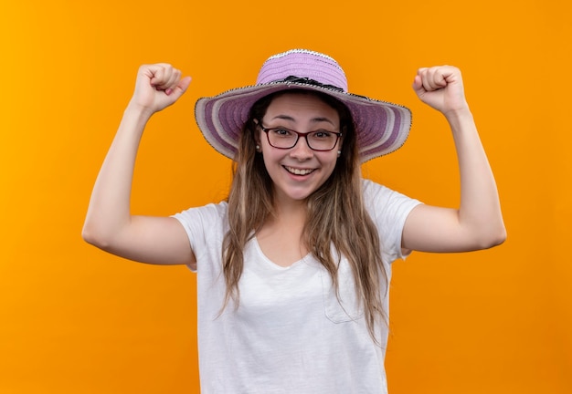 Young woman in white t-shirt  wearing summer hat raising fists happy and excited rejoicing her success  standing over orange wall