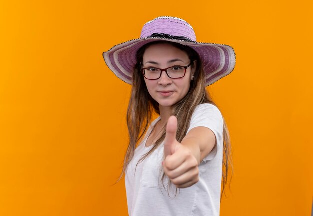 オレンジ色の壁の上に立って親指を元気に笑顔を浮かべて夏の帽子をかぶった白いTシャツの若い女性