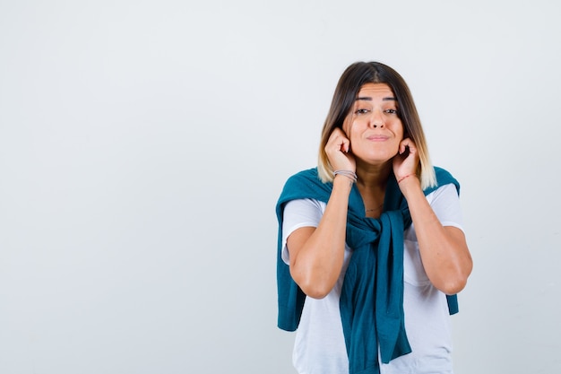 Young woman in white t-shirt with hands near cheeks and looking thoughtful , front view.