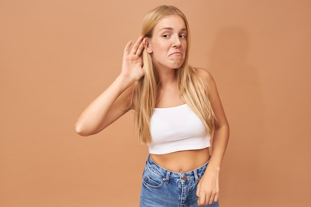 Free photo young woman in white top and blue jeans keeping hand at her ear, listening attentively