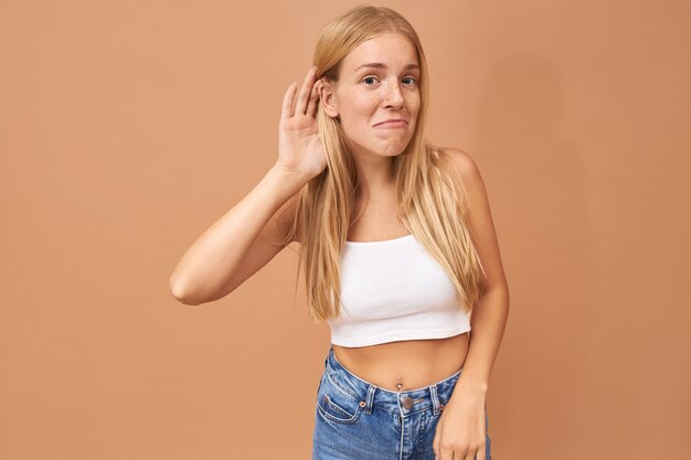 Young woman in white top and blue jeans keeping hand at her ear, listening attentively