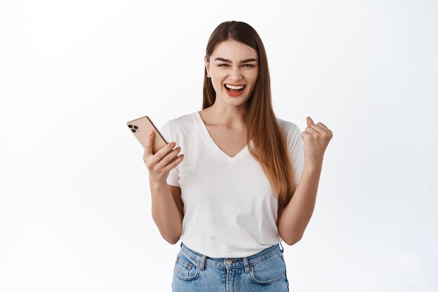 Young woman winning on phone say yes rejoicing and smiling pleased holding smartphone triumphing of app achievement on cellphone standing satisfied against white background