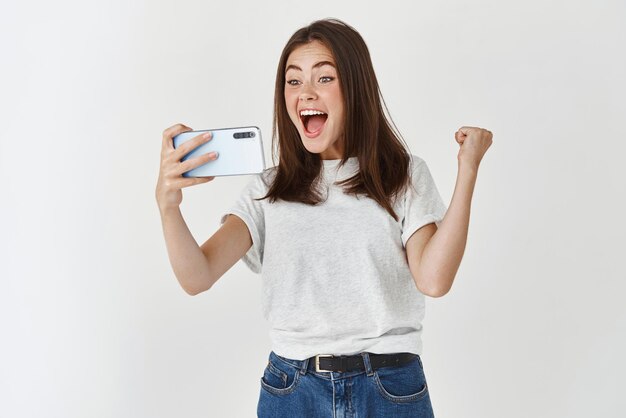 Young woman winning in video game on smartphone scream yes with joy and satisfaction standing over white background