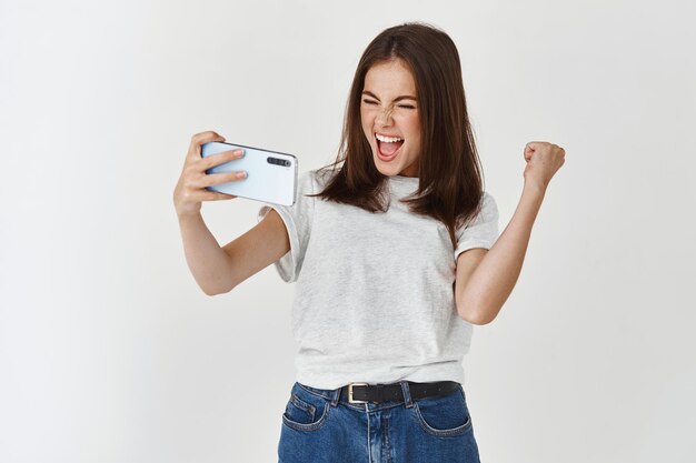 Young woman winning in video game on smartphone, scream yes with joy and satisfaction, standing over white wall