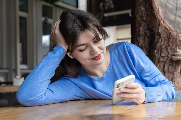 ワイヤレスヘッドホンの若い女性がカフェに座って電話を使用します