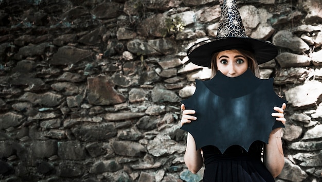 Young woman in witch hat closing mouth with Halloween decoration