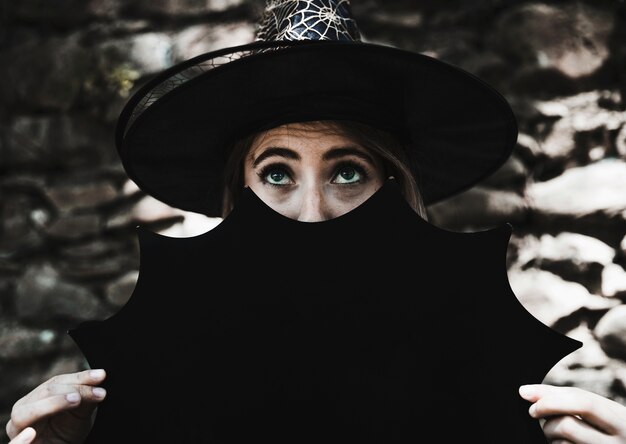 Young woman in witch hat holding Halloween decoration near face