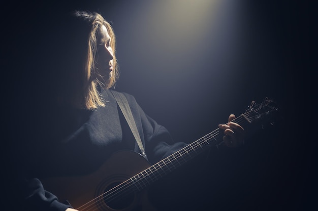 光の光線の下で暗闇の中でアコースティックギターを持つ若い女性