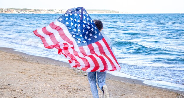 アメリカの国旗を持った若い女性が海を走っています。愛国心と独立記念日のお祝いの概念。