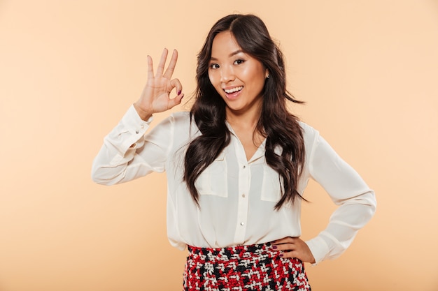 Young woman with asian appearance showing alright gesture being happy and isolated over peach background