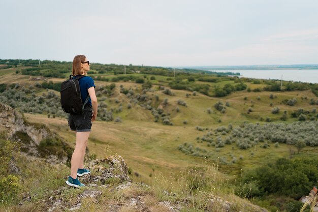 Young woman with backpack standing on cliff's edge and looking to the sky and beautiful nature.