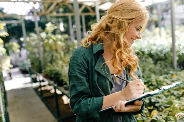 Young woman with beautiful blond hair and gentle smile, dressed in green robe with belt is working in greenhouse