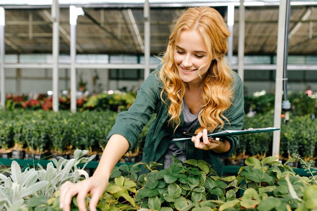 Young woman with beautiful blond hair and gentle smile, dressed in green robe with belt is working in greenhouse