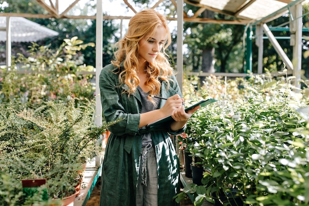 Young woman with beautiful blond hair and gentle smile, dressed in green robe with belt is working in greenhouse