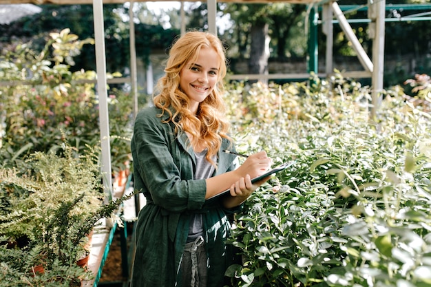美しいブロンドの髪と優しい笑顔の若い女性は、ベルト付きの緑のローブを着て温室で働いています