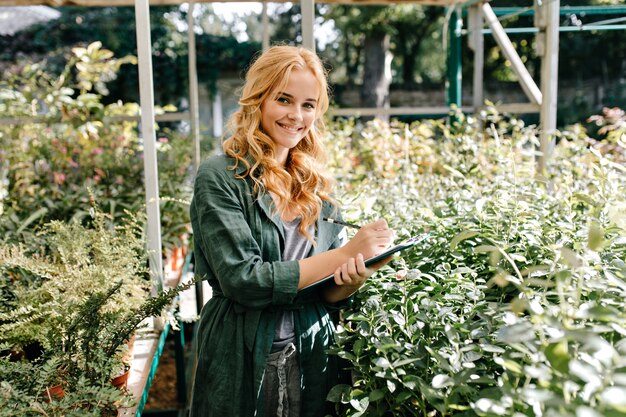 Young woman with beautiful blond hair and gentle smile, dressed in green robe with belt is working in greenhouse