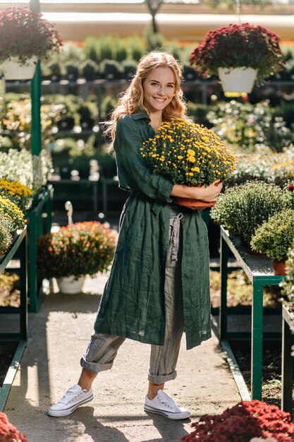 Young woman with beautiful blond hair and gentle smile, dressed in green robe with belt is working in greenhouse